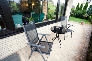 Un patio con dos sillas, una mesa, un muro de ladrillo y un amplio ventanal con vistas a un jardín.