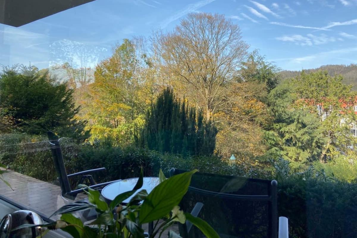 Une vue depuis un balcon, avec une table et des chaises en verre, donnant sur un jardin où l’on peut admirer différentes espèces d’arbres et un bâtiment situé au loin.