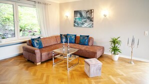 Ein modernes Wohnzimmer mit einem braunen Ledersofa, einem Glastisch und einem weißen Hocker. Ein großes Fenster mit weißen Vorhängen, eine Zimmerpflanze und ein an der Wand befestigtes Kunstwerk sind ebenfalls zu sehen.