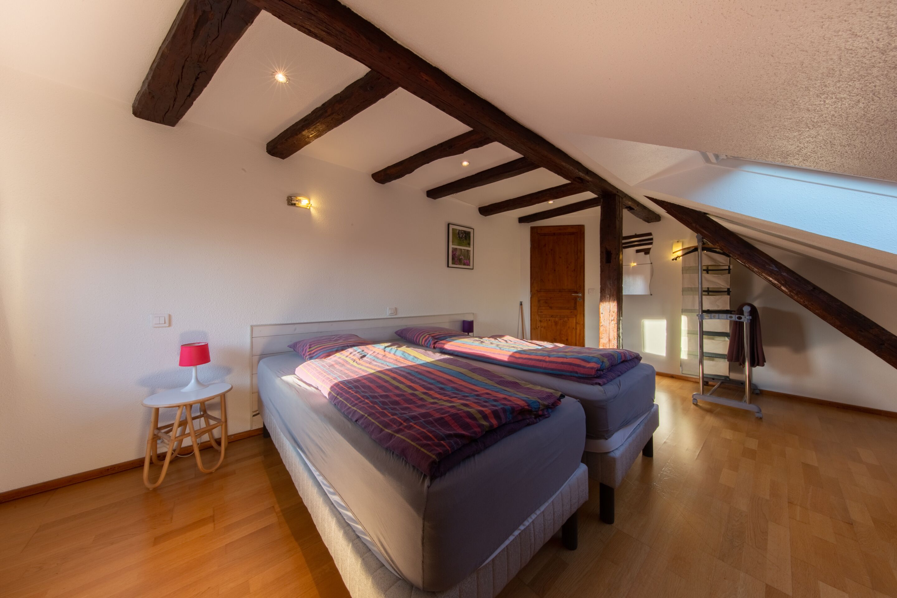 Un dormitorio con techo inclinado, vigas de madera, una cama con una colcha colorida, una mesita de noche con una lámpara y una ventana con vistas.