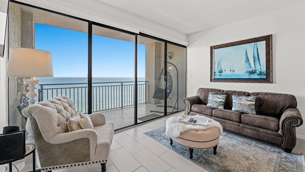 A spacious living room with a large sliding glass door leading to a balcony with a view of the ocean. There is a brown leather sofa, a beige armchair, and a small ottoman with a white furry throw. A painting of sailboats hangs on the wall.