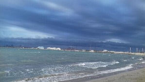 Udsigt over kysten med strand, hav og fjerne bygninger under en overskyet himmel.