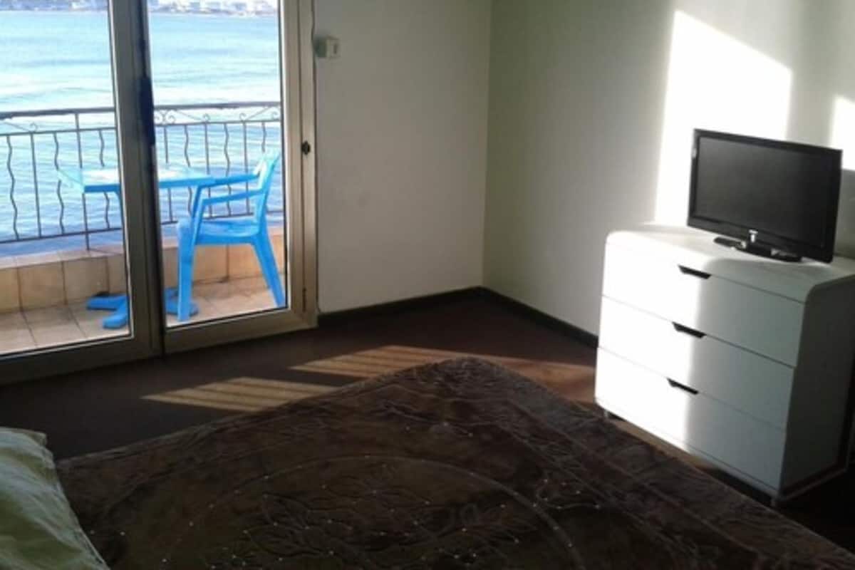 A hotel room with a bed, a TV on a dresser, a balcony with a view of the sea, and a blue chair.