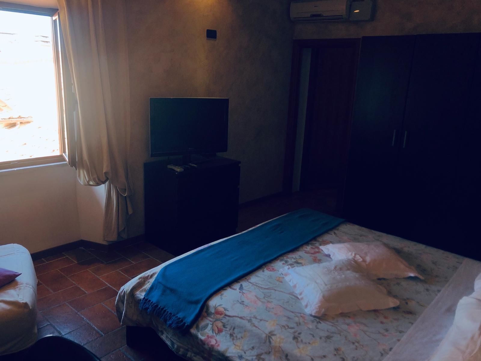Une chambre à coucher avec un lit, un téléviseur posé sur un meuble, une armoire et une fenêtre avec des rideaux.