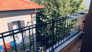 Een balkon met betegelde vloer, een metalen hekwerk en uitzicht op een gebouw met een rood dak en een raam met luiken.