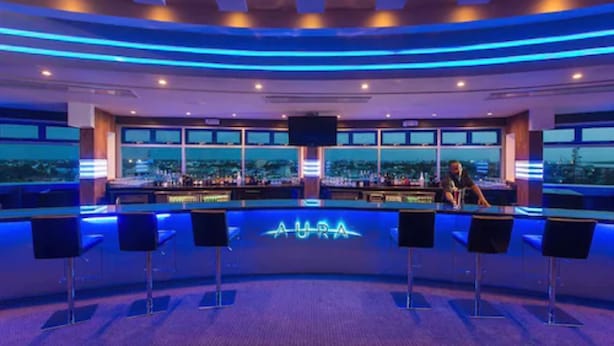 Un'area bar con un neon che riporta la scritta "AURA" e diversi sgabelli da bar.