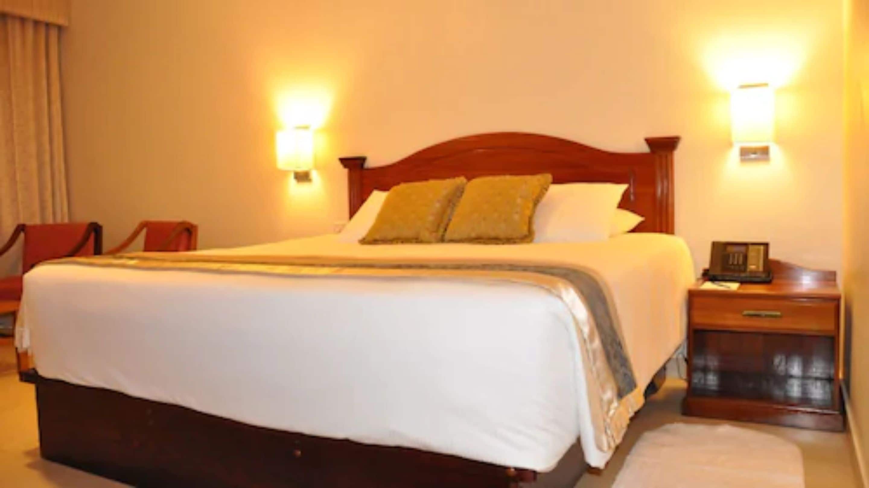 Habitación de hotel con una cama grande, dos mesitas de noche, una silla y lámparas de pared.