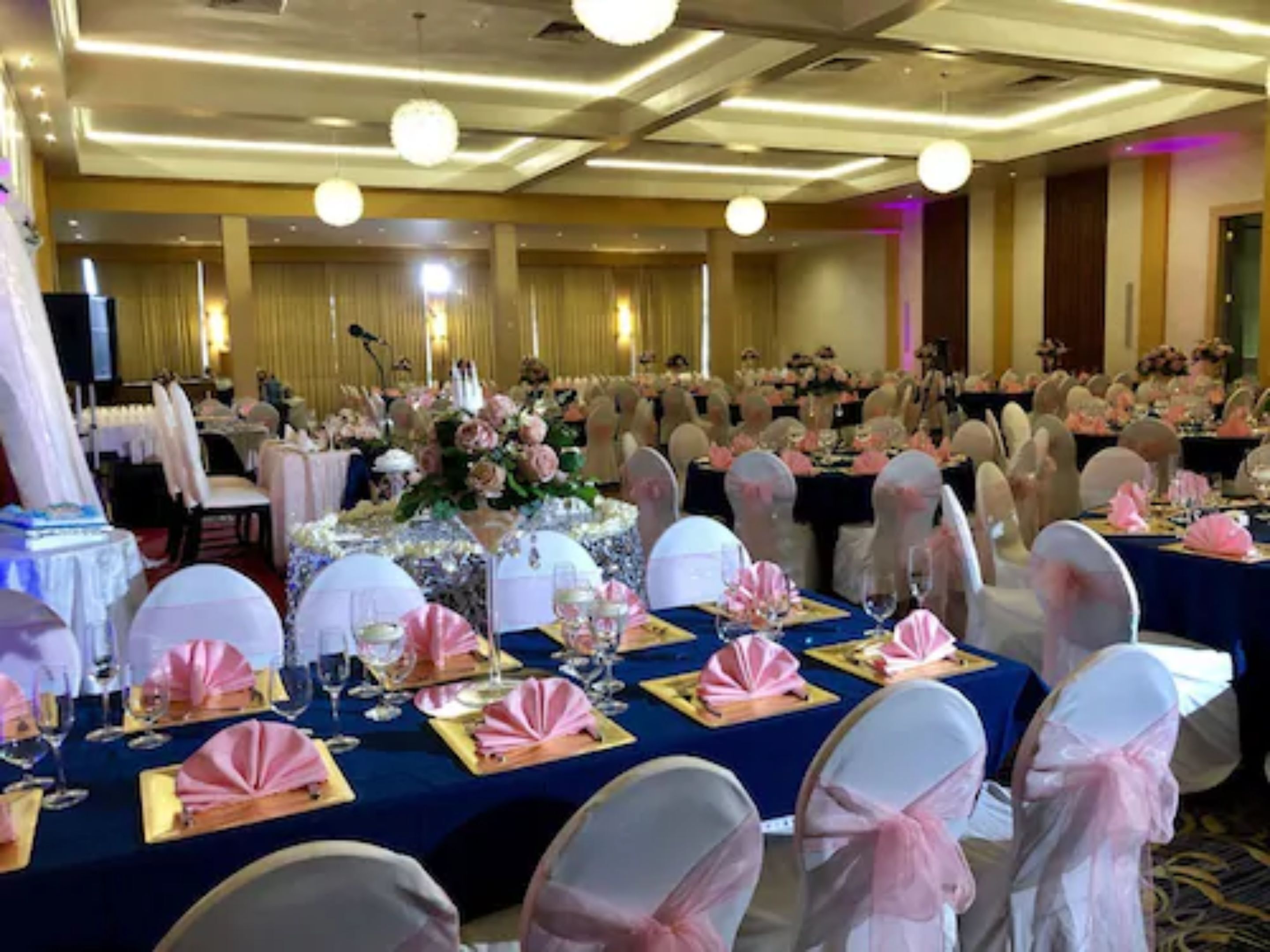 A banquet hall with round tables set for a formal event, featuring white chairs with pink sashes and blue tablecloths.