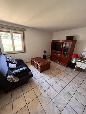 A living room with a sofa, a wooden cabinet, a television, and a window. - Chambres des Arrys (Bas-en-Basset)