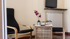 Una habitación con un sofá negro, una silla de madera, una mesita con un jarrón de flores moradas, una bandeja con aperitivos y un televisor en la pared.