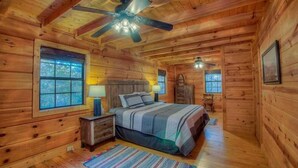 A cozy log cabin bedroom with a bed, two bedside tables, a ceiling fan, and large windows. - Woodentops- Hot Tub, Pet Friendly, Toccoa River Access (Mineral Bluff)