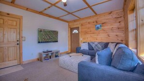 A cozy living room with a wooden ceiling, a grey sofa, a television mounted on the wall, and a wooden door. - Woodentops- Hot Tub, Pet Friendly, Toccoa River Access (Mineral Bluff)