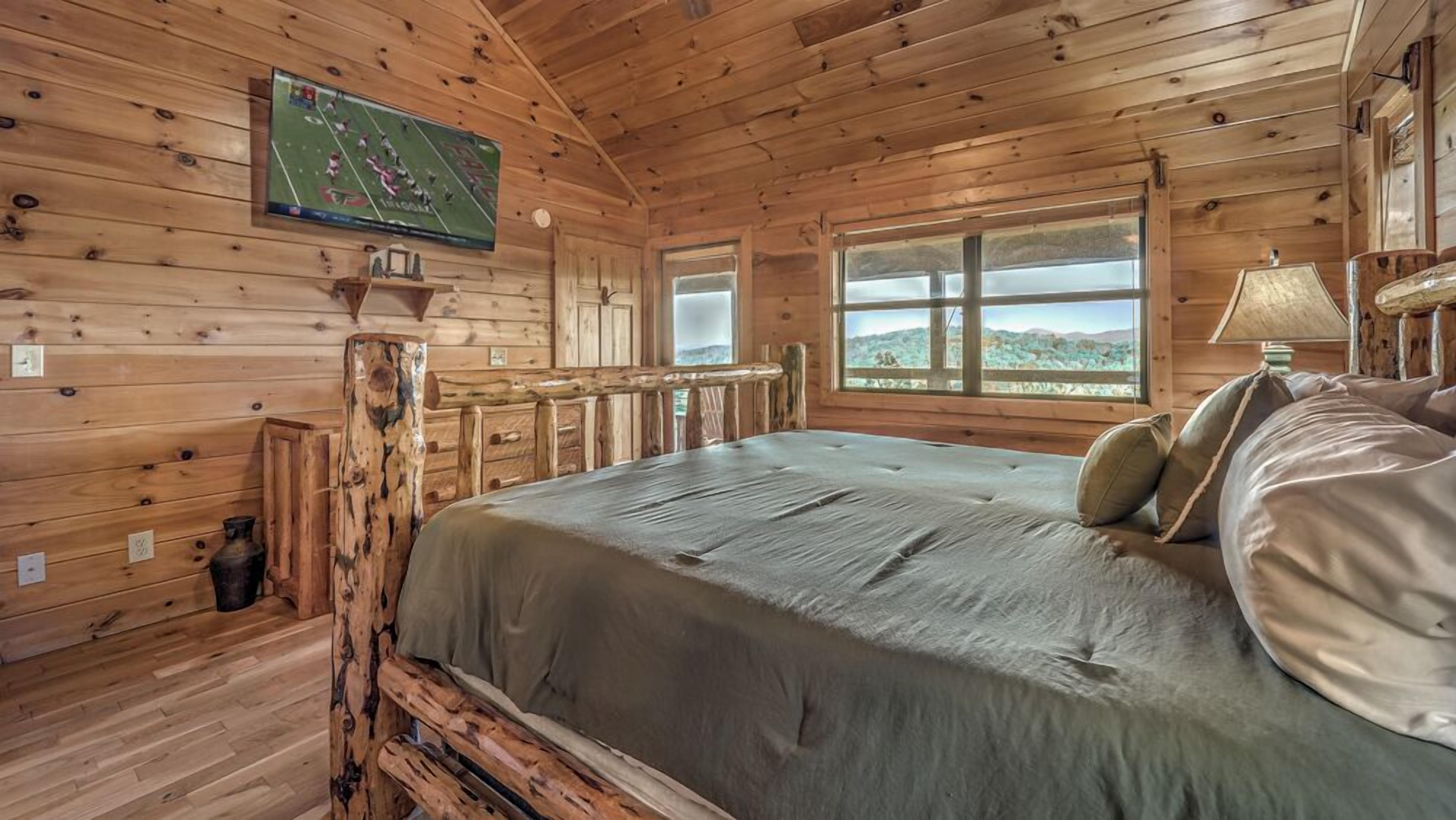 A wooden cabin room with a bed, a television mounted on the wall, and large windows offering a view of the outdoors.