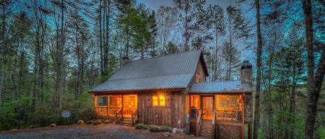 A rustic cabin with a metal roof, wooden exterior, and a stone chimney. It features a porch with a railing and a satellite dish.