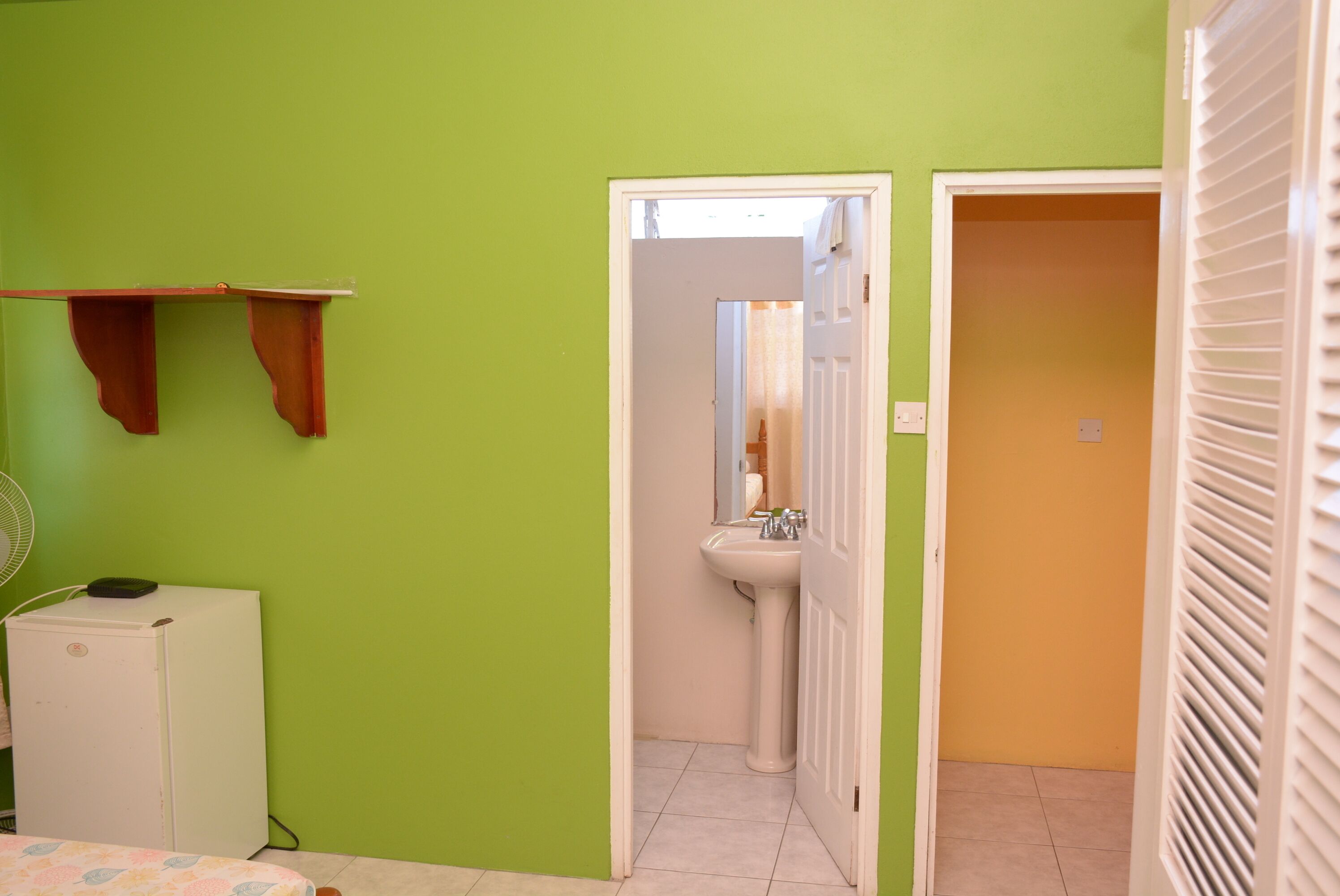 Une pièce avec un mur vert, un réfrigérateur blanc, un lit recouvert d’une couvre-lit à motifs floraux, un lavabo blanc et une porte donnant sur une pièce orange.