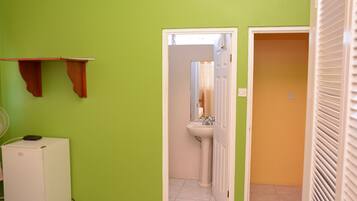 Una habitación con una pared verde, un refrigerador blanco, una cama con edredón floral, un lavamanos blanco y una puerta que da a una habitación naranja.