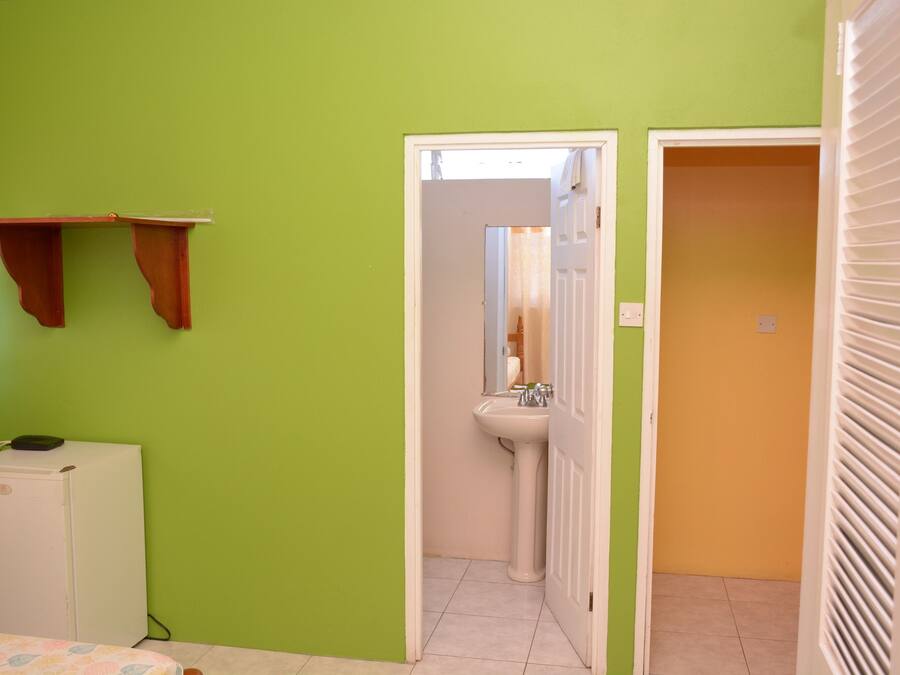 Ein Zimmer mit grüner Wand, einem weißen Kühlschrank, einem Bett mit floralem Überwurf, einem weißen Waschbecken und einer Tür zu einem orangefarbenen Raum.