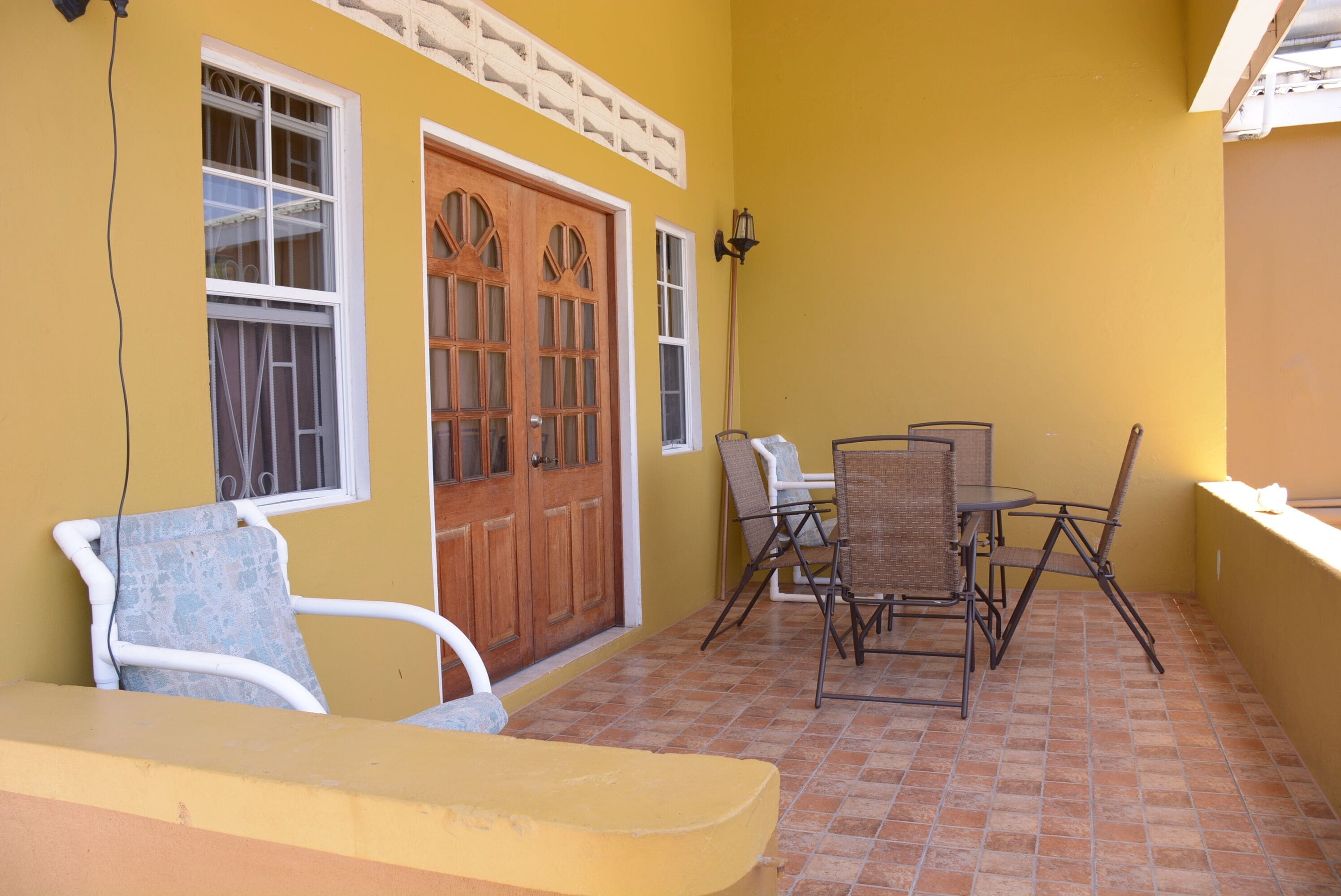 A yellow building with a wooden door, white-framed windows, and a tiled patio area with a table and chairs.