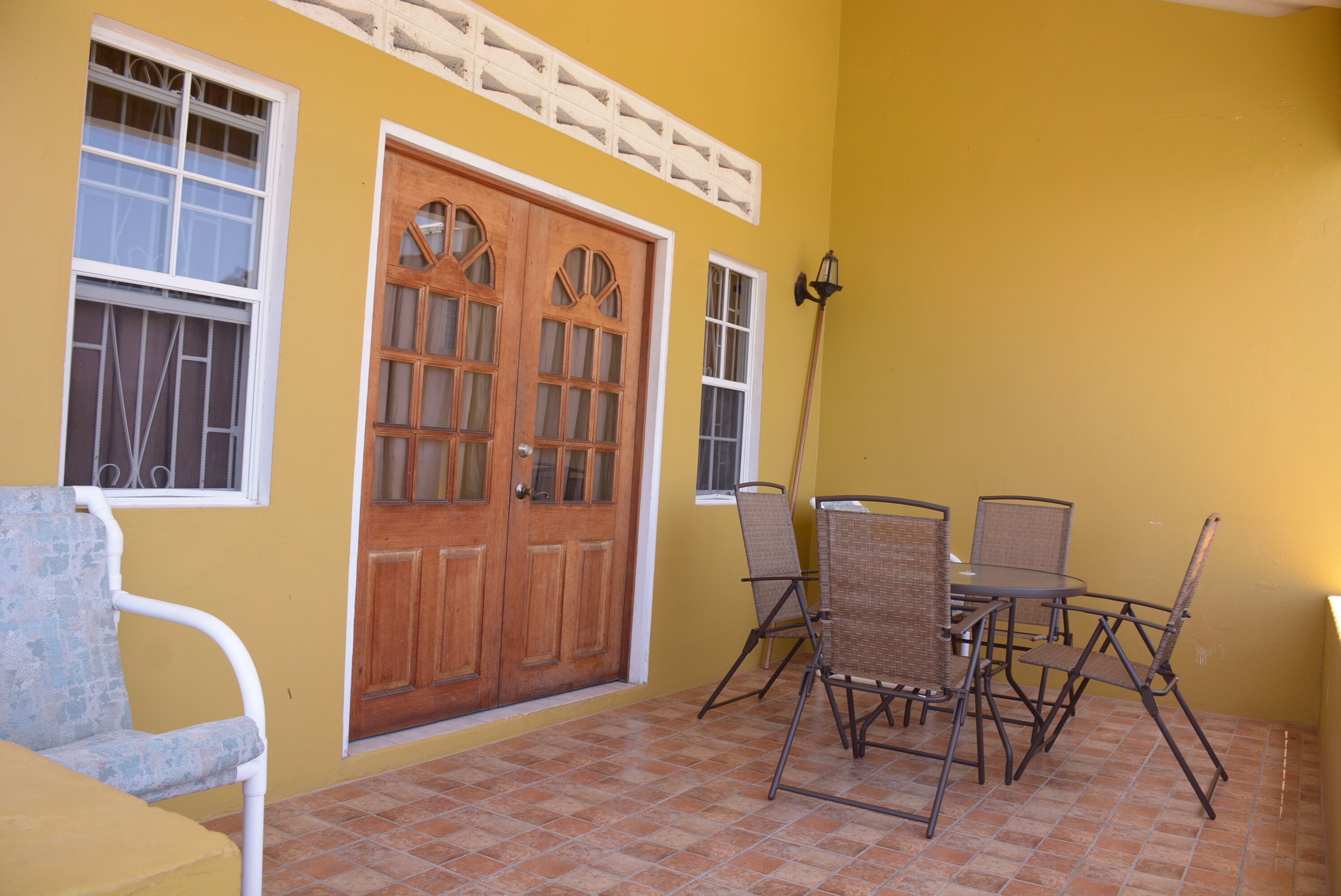 A yellow building with a wooden door, white-framed windows, and a tiled patio with metal chairs and a table.