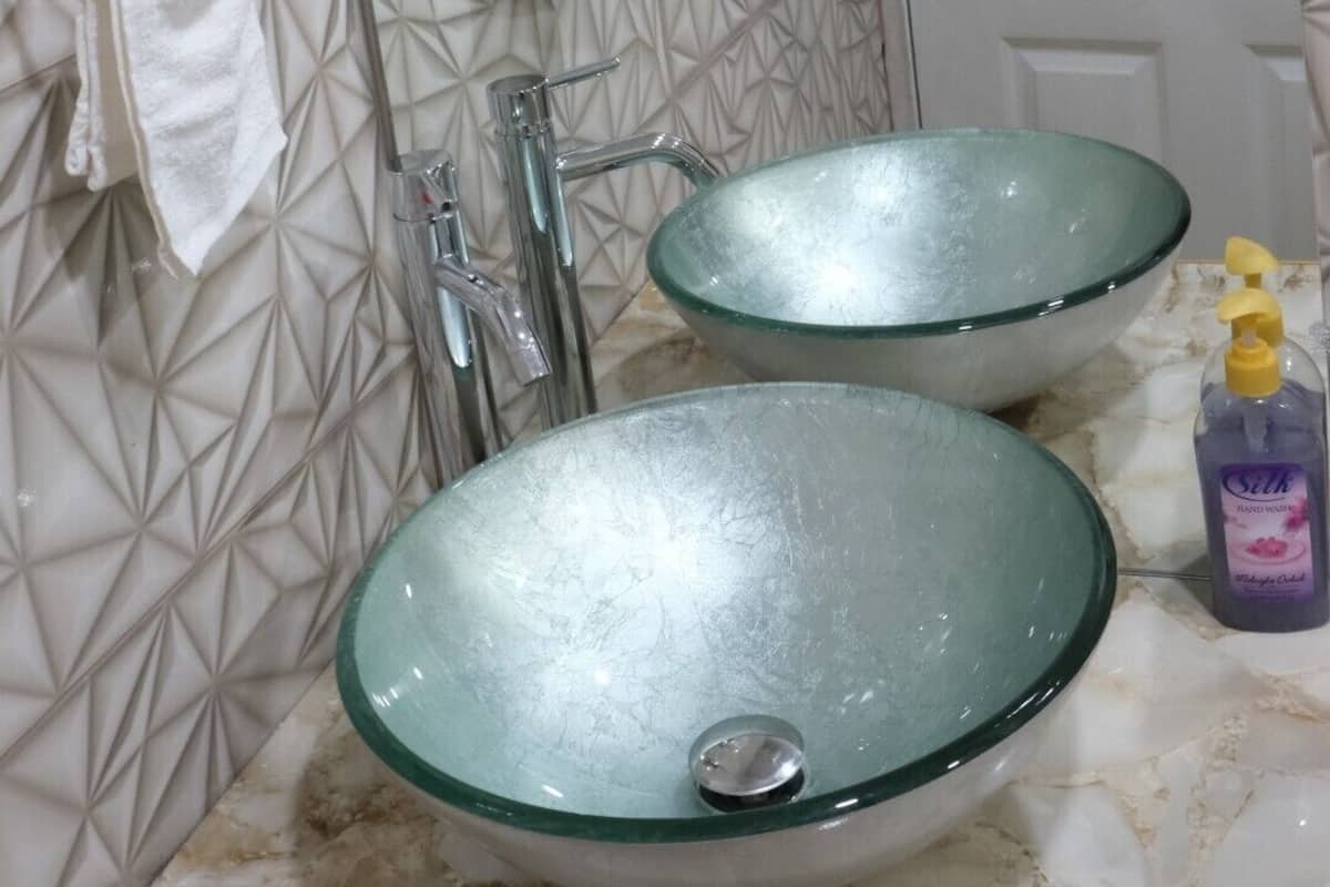 A bathroom with a geometric patterned wall, two glass sinks, a mirror, a towel rack, and a door.