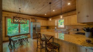 A wooden cabin kitchen with a large island, bar stools, and a view of the forest through the windows. - Sleepy Bear Hideaway - Close to Train, Mountain Views, Hot Tub (Blue Ridge)