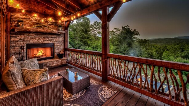 A cozy outdoor seating area with a stone fireplace, wicker furniture, and a view of the surrounding forest. - Sleepy Bear Hideaway - Close to Train, Mountain Views, Hot Tub (Blue Ridge)