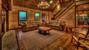 A cozy log cabin interior with a fireplace, two patterned sofas, a coffee table, and a staircase leading to a balcony. - Sleepy Bear Hideaway - Close to Train, Mountain Views, Hot Tub (Blue Ridge)