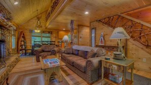 A cozy log cabin interior with a fireplace, leather sofas, a coffee table, and a bookshelf. - Riverwalk- to Toccoa River | Screened Porch | Pet Friendly | Fenced Yard (Blue Ridge)
