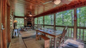 A rustic cabin with a stone fireplace, wooden furniture, and large windows offering a forest view. - Riverwalk- to Toccoa River | Screened Porch | Pet Friendly | Fenced Yard (Blue Ridge)