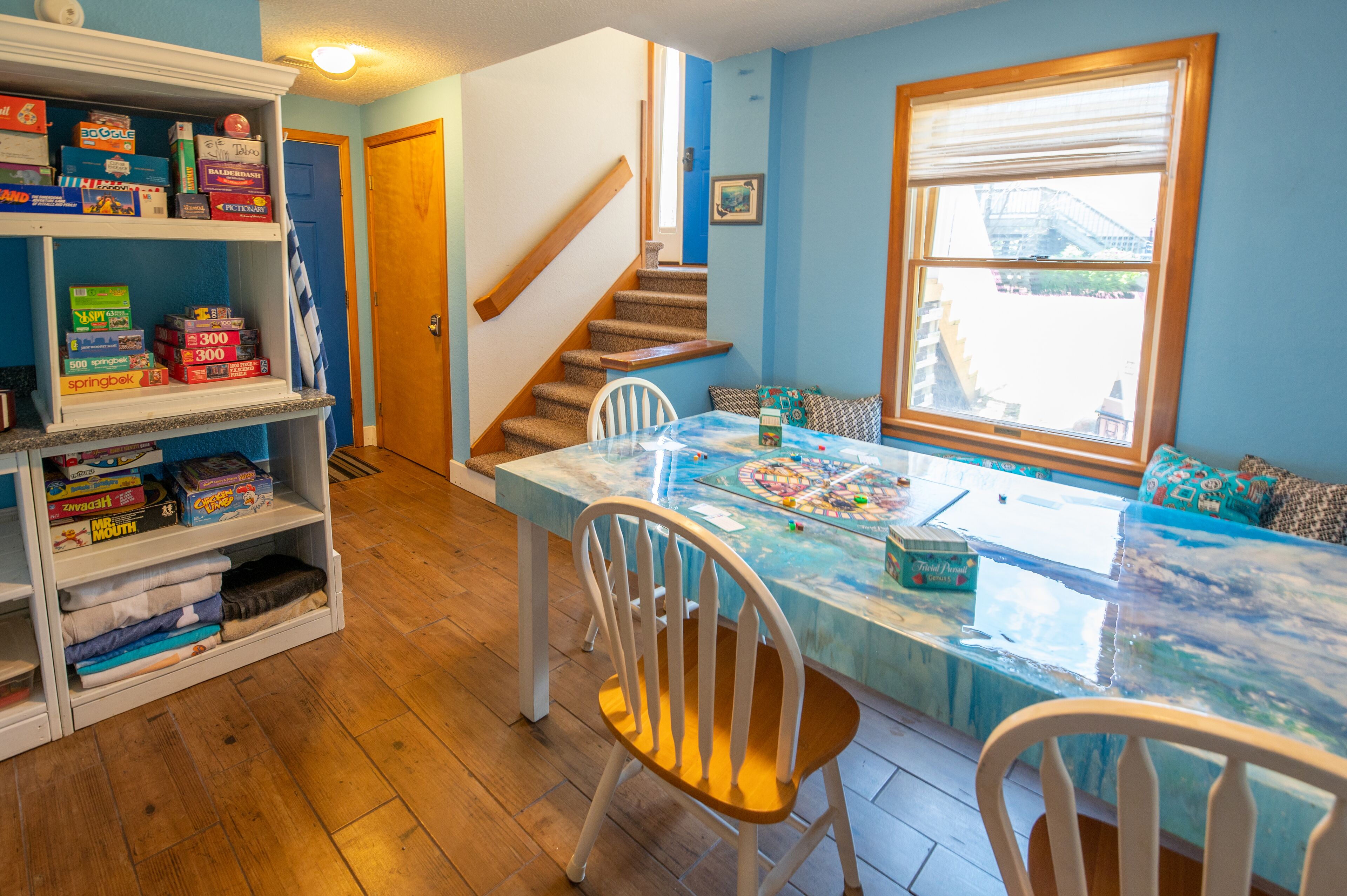 Una habitación con piso de madera, una mesa de comedor con sillas, una estantería llena de juegos de mesa, una escalera y una ventana con vistas a edificios.