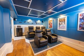 Sala de cinema com paredes azuis, piso de madeira e quatro poltronas reclináveis.