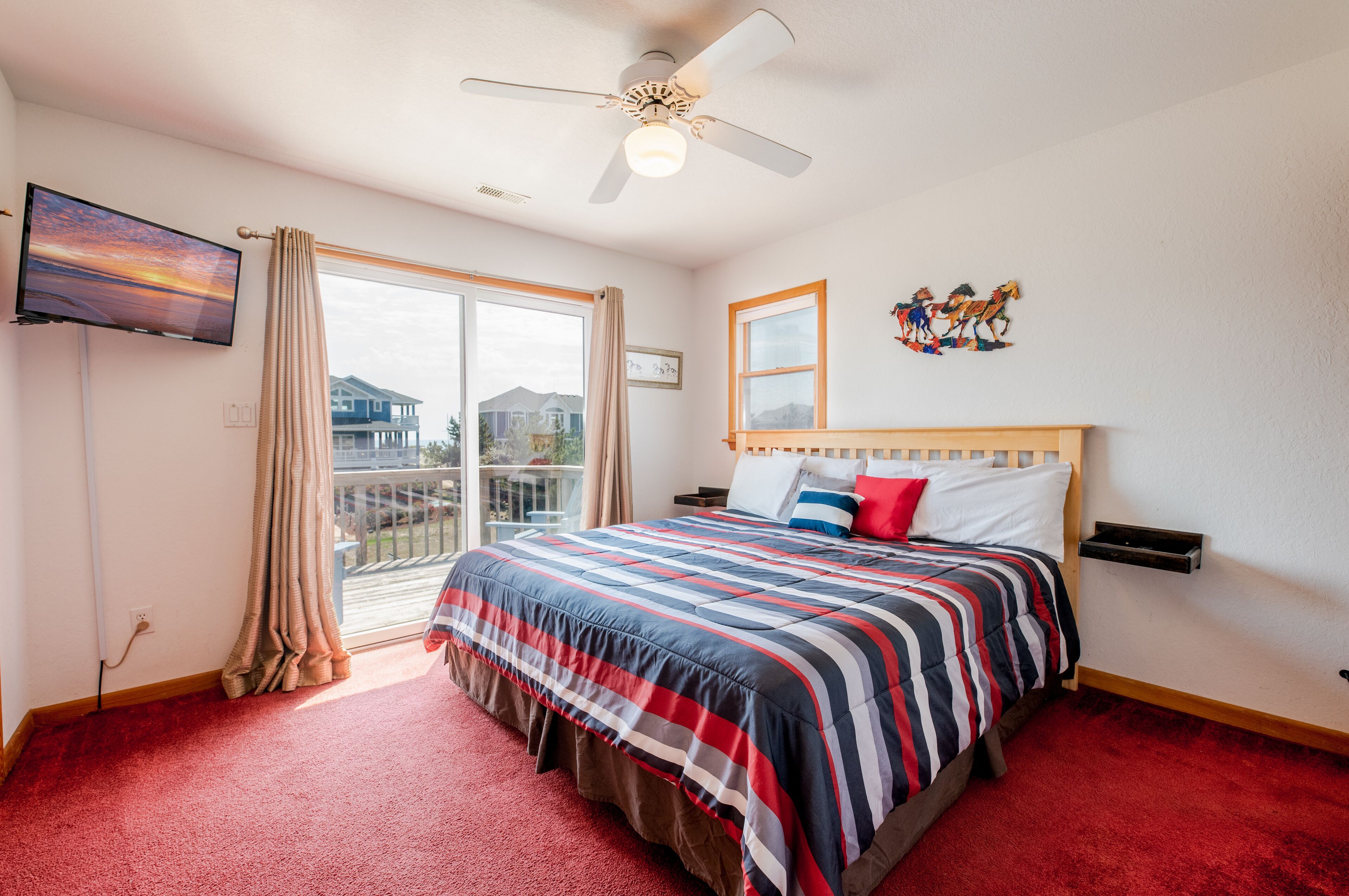 A hotel room with a large bed, a ceiling fan, a TV mounted on the wall, a window with curtains, and a balcony with a view of houses.