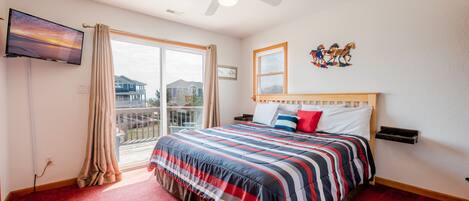 A hotel room with a large bed, a ceiling fan, a TV mounted on the wall, a window with curtains, and a balcony with a view of houses.