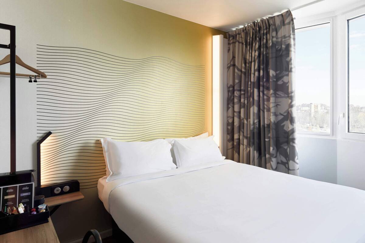 A hotel room with a large bed, a bedside table with a lamp, and a view of the city through a window with curtains.