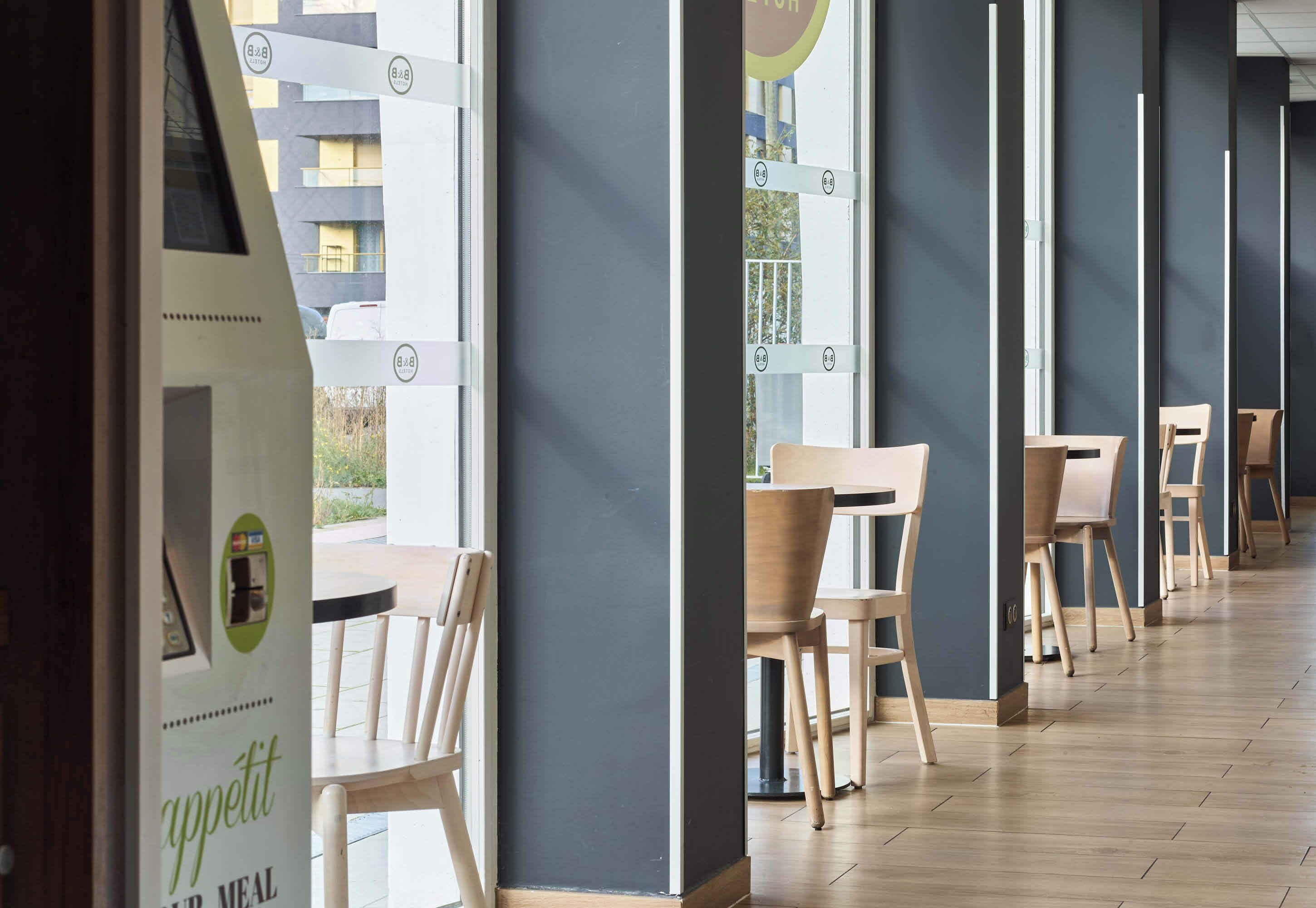 Un comedor moderno con sillas y mesas de madera, ventanas amplias y vistas a un edificio.