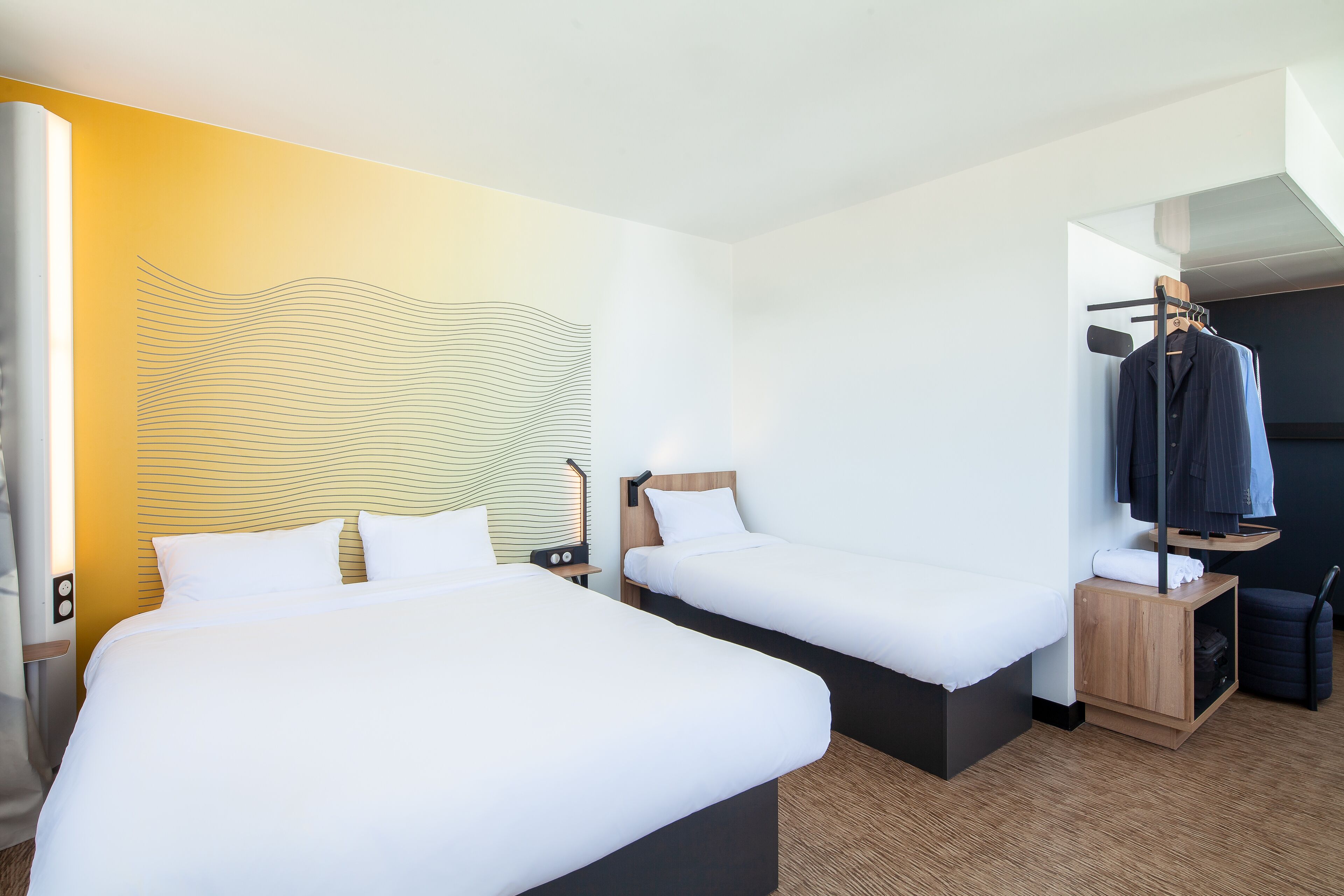 Una habitación de hotel con dos camas, una mesita de noche de madera, un perchero y una lámpara fijada a la pared.