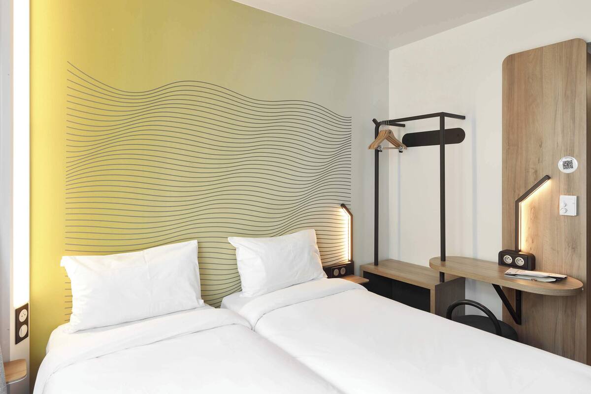 A hotel room with a bed, bedside tables, a wooden headboard, a wall-mounted coat rack, and a desk with a chair.