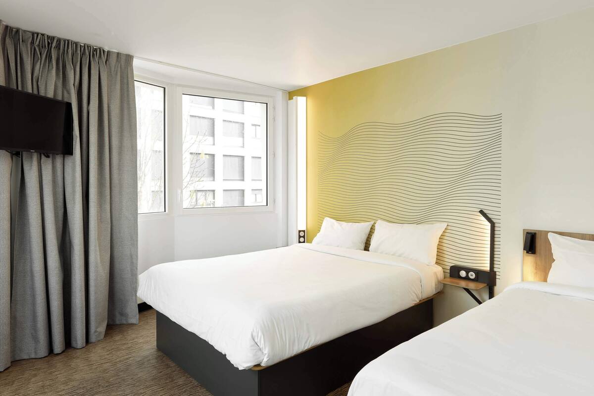 A hotel room with a large bed, a bedside table with a lamp, a flat-screen TV mounted on the wall, and a window with curtains.