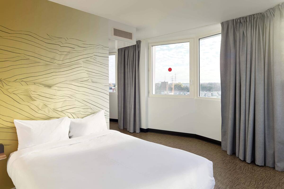 A hotel room with a large bed, a view of the city through a window, and a wall with a textured design.