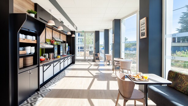 A modern cafe with a long counter, seating area, and large windows. - B&B HOTEL MASSY Gare TGV (Massy)