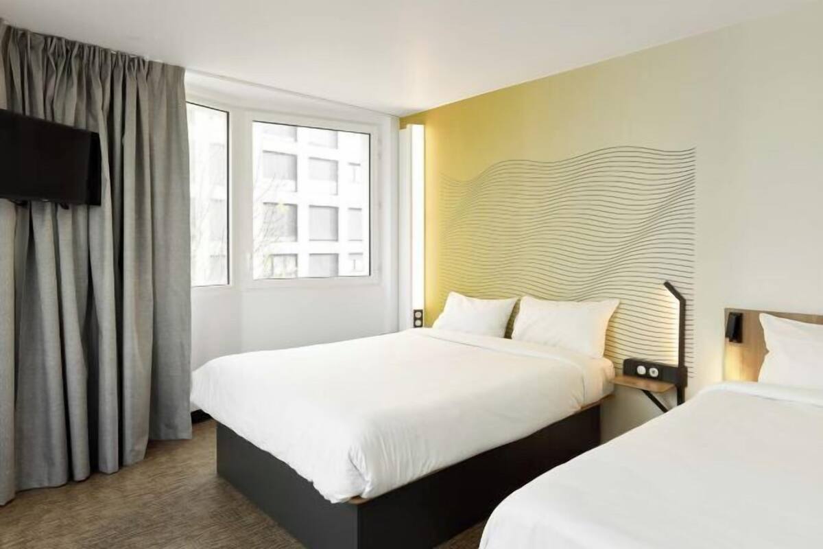 A hotel room with two beds, a TV, a window with curtains, and a unique wall design.