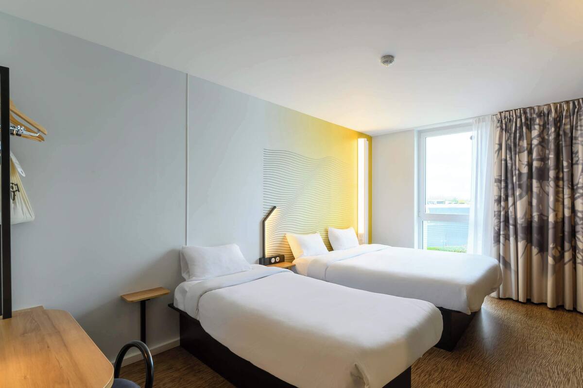A hotel room with two beds, a wooden table, a small desk, and a window with curtains.