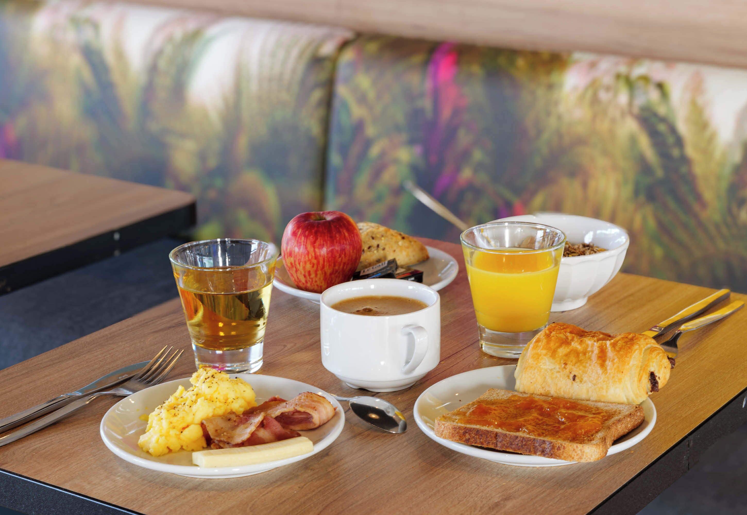 A breakfast setting with plates of eggs, bacon, toast, and a croissant, accompanied by a glass of orange juice and a cup of coffee.