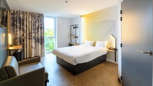 Una habitación de hotel moderna con una cama grande, un escritorio y una silla. Cuenta con una ventana con cortinas, una lámpara de pared y una puerta.