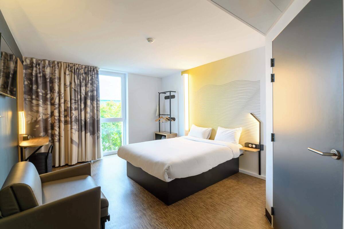 A modern hotel room with a large bed, a desk, and a chair. There is a window with curtains, a wall-mounted lamp, and a door.