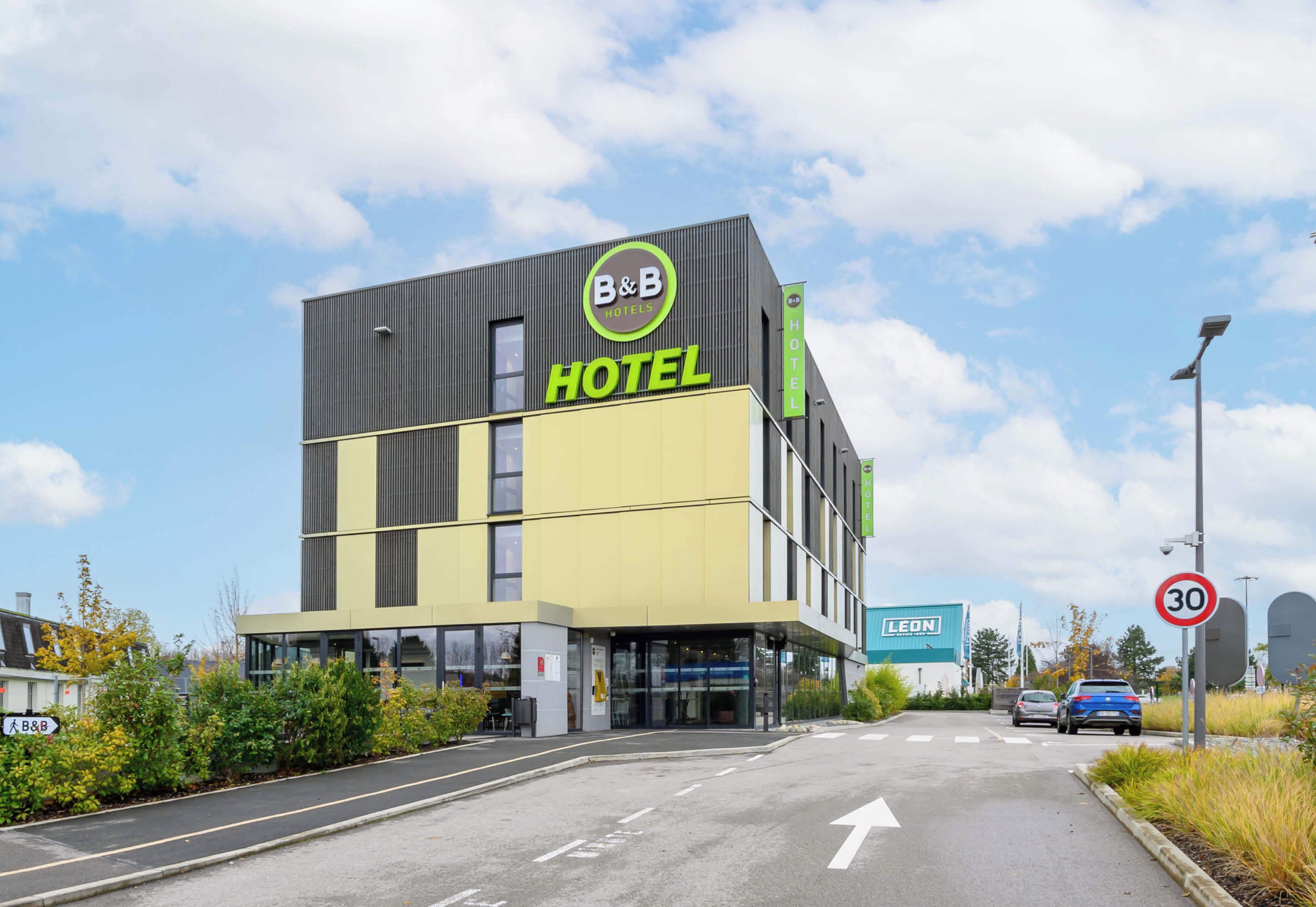A modern B&B hotel with a prominent sign, a clear road in front, and a speed limit sign.