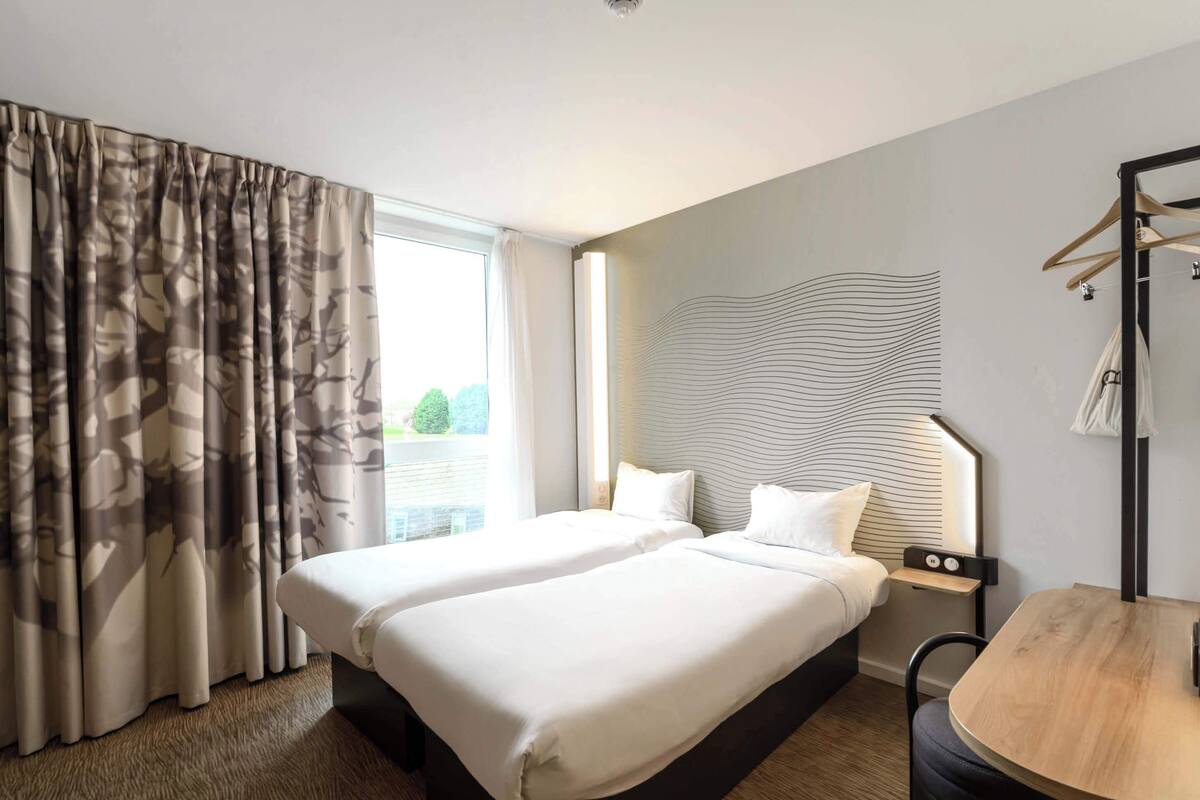 A hotel room with two beds, a wooden table, a chair, a coat rack, and a view of greenery outside.