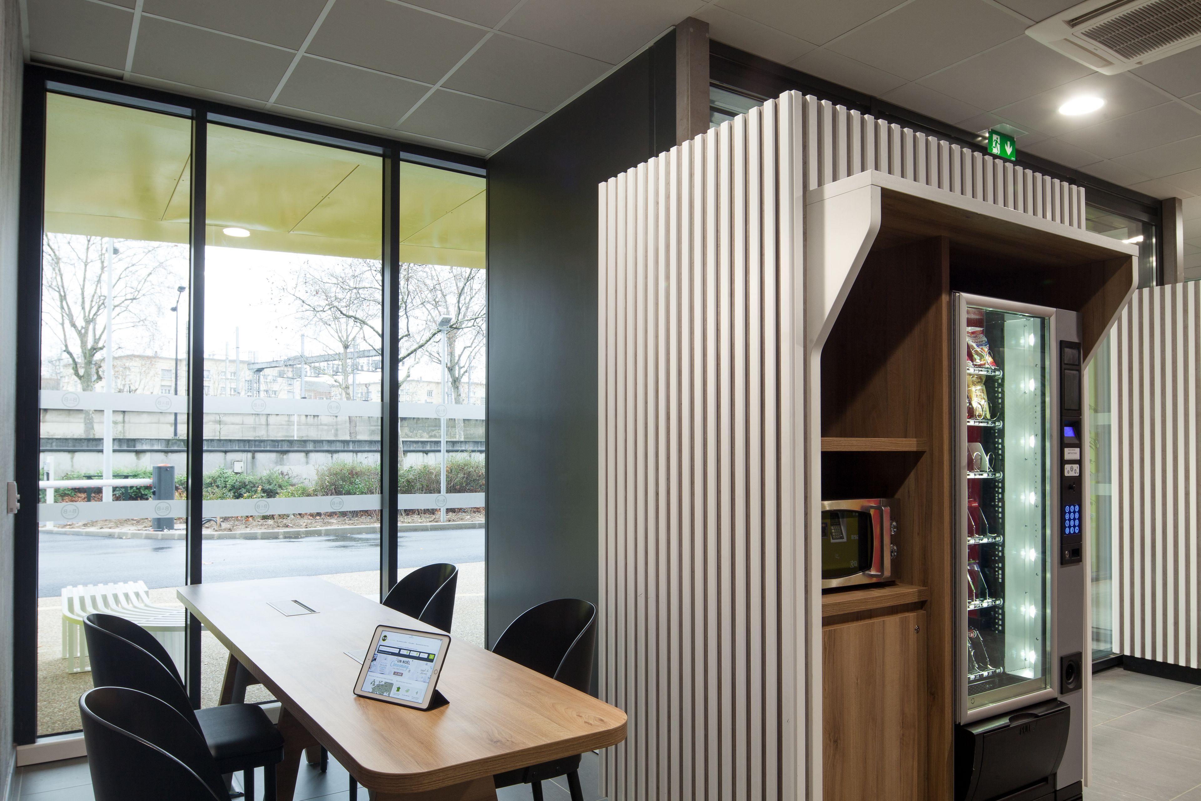 Ein modernes Interieur mit einem Holztisch und -stühlen, einem Verkaufsautomaten und Blick ins Freie durch große Fenster.