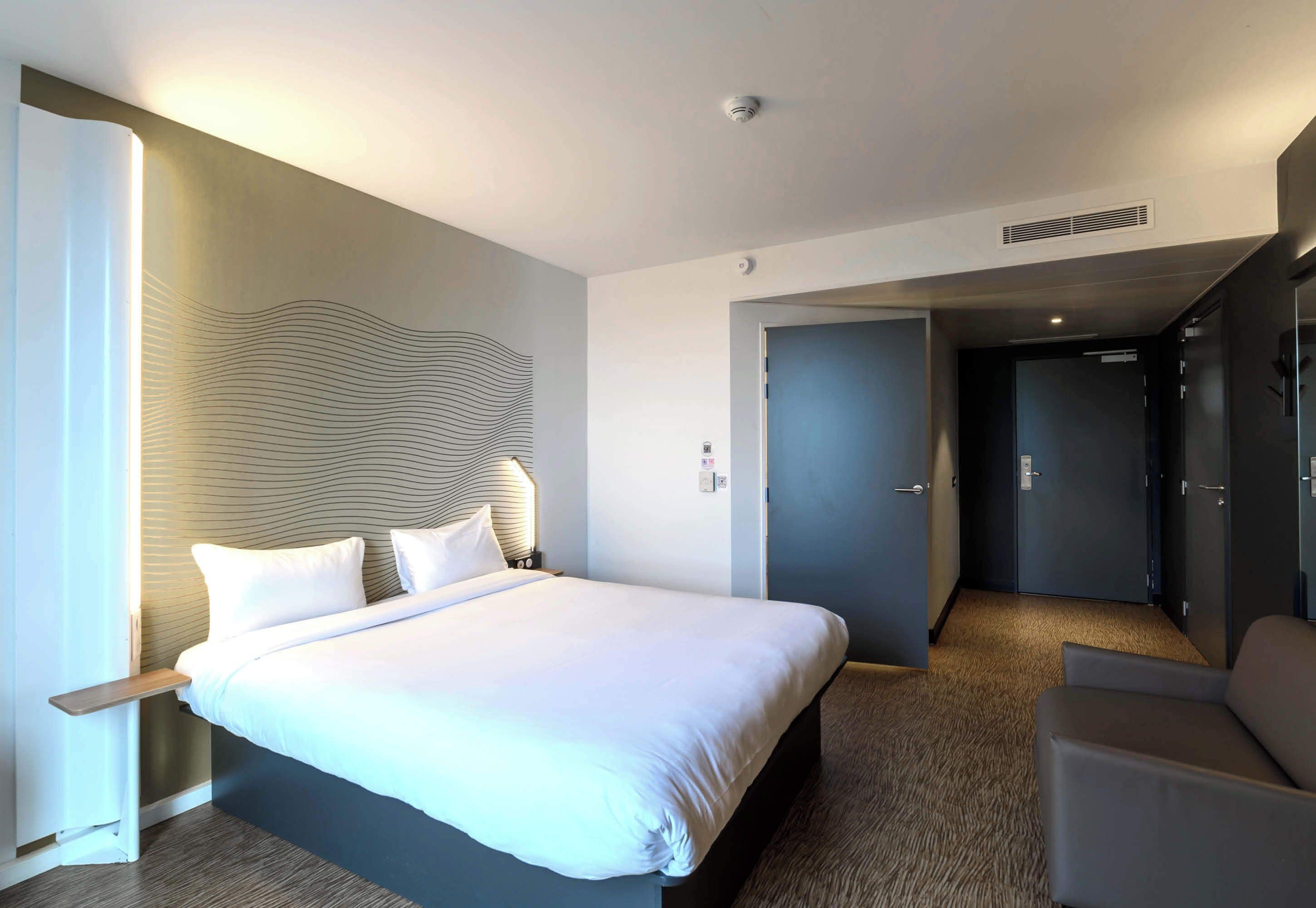 A modern hotel room with a large bed, a bedside table, a chair, and a door leading to a hallway.