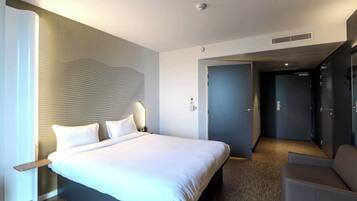 Une chambre d’hôtel moderne, dotée d’un grand lit, d’une table de chevet, d’une chaise et d’une porte donnant sur un couloir.
